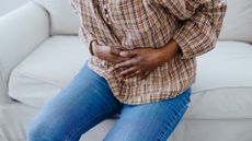 Close-up of a woman holding her hands over her pelvis in pain. She is wearing a brown checked shirt and blue denim jeans and is sat on a white couch.
