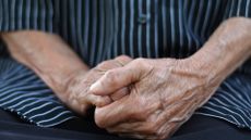 Close-up of the hands of an elderly person that are crossed together over their lap. They are wearing a shirt with black and light blue stripes.